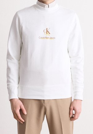Biała bluza z okrągłym dekoldem z logo Calvin Klein Jeans w beżowym i brązowym kolorze, noszona na paskowaną koszulę z kołnierzykiem, zestawiona z beżowymi spodniami.