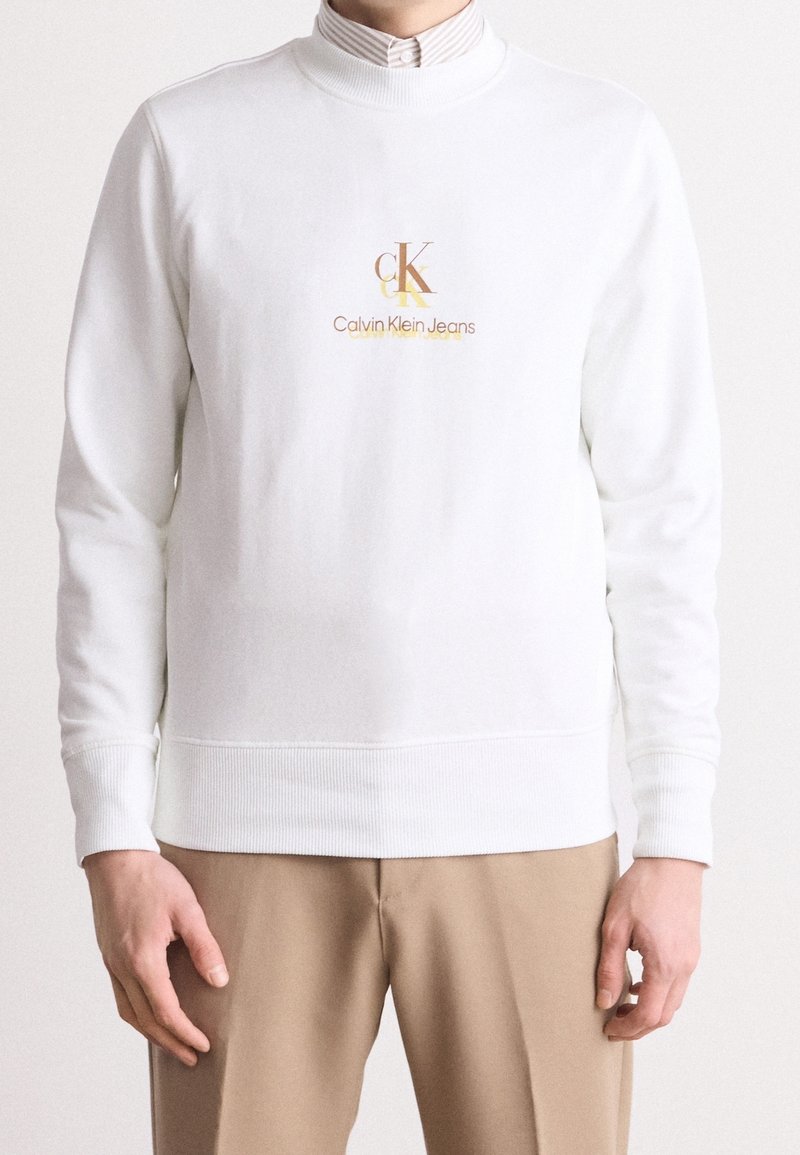 Sweat-shirt blanc à col rond avec logo Calvin Klein Jeans en beige et marron, porté sur une chemise rayée à col, associé à un pantalon beige.