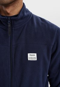 Chaqueta de forro polar en color azul marino con cuello alto y cierre de cremallera. Incluye una etiqueta blanca con texto negro cosida en el pecho. Textura suave.