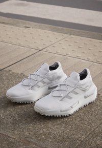 Pantofi sport albi cu o textură din tricot, având accente gri și un design cu trei dungi. Talpa groasă cu un fund aderent și texturat.