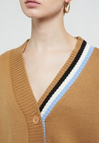 Brauner gestrickter Cardigan mit V-Ausschnitt, einer einzelnen Knopfleiste und mehrfarbigen Streifen in Blau, Weiß und Schwarz entlang des Ausschnitts.