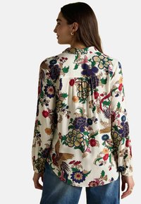 Woman facing away wearing a white long-sleeve blouse with colorful floral and bird patterns and blue jeans.