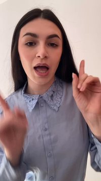 Noor naine, kellel on pikad tumedad juuksed ja kes teeb sõrmega ülespoole viitavaid žeste, kannab sinist särki lillemustriga kraega, taust on ühtlane.