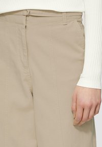 Pantalons larges beiges avec une texture lisse, dotés d'une taille droite et de détails de coutures visibles à l'avant.