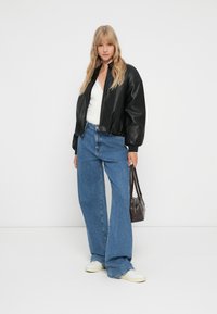 Blonde Frau in schwarzer Lederjacke, weißem Oberteil, blauen weiten Jeans und weißen Sneakers, die eine dunkelbraune Handtasche vor einem weißen Hintergrund hält.