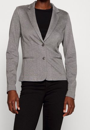 Donna che indossa un blazer grigio aderente con due bottoni sopra una maglia nera e pantaloni neri, in piedi davanti a uno sfondo chiaro e uniforme.
