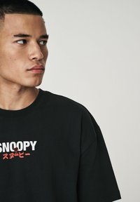 Sorte bomulds T-shirt med "SNOOPY" i fed hvid og rød tekst samt en lille tegneserie-grafik. Korte ærmer og rund halsudskæring.