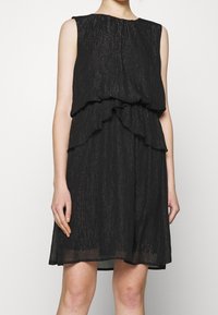 Schwarzes ärmelloses Kleid mit einem gestuften Rüschendesign, aus strukturiertem Stoff mit subtiler Glanzoptik, knielang und locker geschnitten.