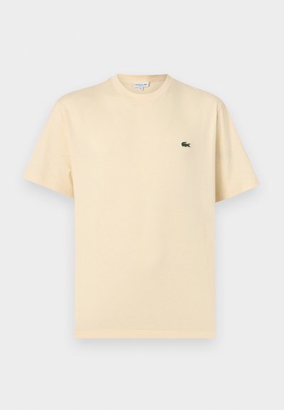 T-shirt beige à manches courtes et col rond, avec un petit logo crocodile vert sur la poitrine gauche, présenté sur un fond blanc.