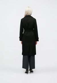 Cappotto trench in lana nera con vita cintata, maniche lunghe e dettagli con polsini abbottonati, abbinato a pantaloni grigi a gamba larga e tacchi neri.