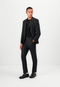 Schwarzer Anzug mit taillierter Passform, einreihige Jacke, passende Hose, getragen über einem schwarzen Oberteil, kombiniert mit schwarzen Schuhen, akzentuiert durch eine goldene Kette.