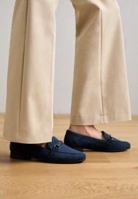 Mocasines de ante color azul marino con punta redondeada, pequeño tacón y detalle de hardware plateado en la parte superior. Combinados con pantalones de pierna ancha beige.
