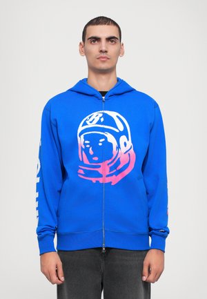 Fato de treino azul com fecho éclair, feito de algodão, com um gráfico de um astronauta em rosa e branco na frente e texto na manga esquerda.