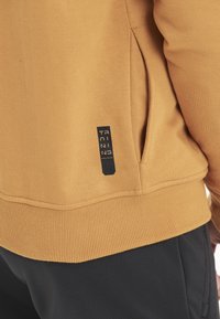 Sennepsgul sweatshirt med ribbet kant og manchetter, sidelomme og et blåt logo-patch med teksten "TRAINING ENDURANCE."