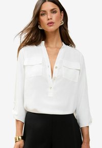 Camicia bianca con scollatura a V profonda, due tasche sul petto e maniche arrotolate. Tessuto liscio, abbinata a pantaloni neri.