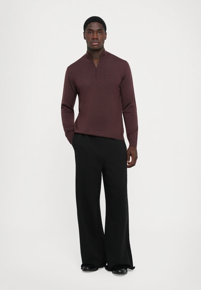Pull en molleton bordeaux à manches longues avec un col zippé jusqu'au quart, associé à un pantalon large noir et des chaussures noires. Texture lisse, design ajusté.