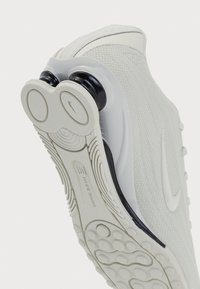 Nike Shox kedas ar teksturētu balto sietu augšdaļu, diviem cilindriskiem melniem papēžiem, gumijas zoli un izteiksmīgu zīmoljūtas vidusdaļā.