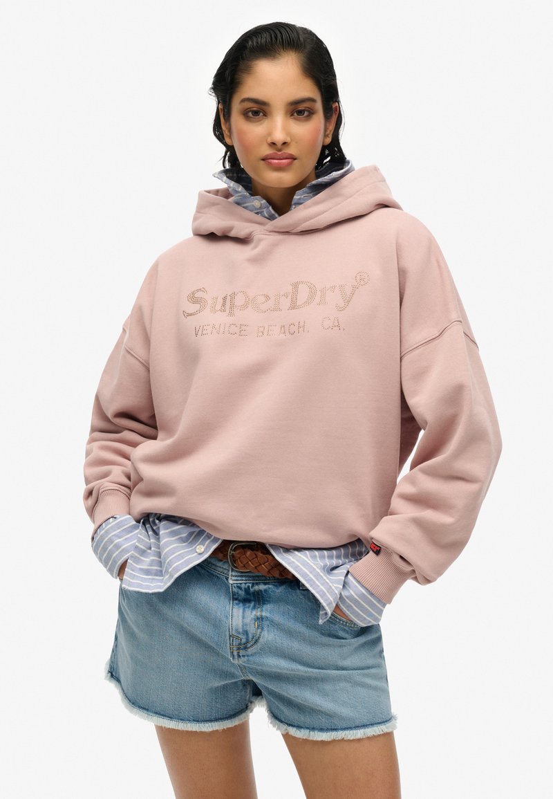 Felpa rosa con logo "SuperDry", camicia a righe blu chiaro sotto, pantaloncini di jeans e una cintura intrecciata marrone. Tessuto morbido e casual, con una vestibilità rilassata.