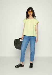 Mulher vestindo blusa amarela, jeans azuis, tamancos pretos e óculos de sol com tonalidade laranja, segurando uma bolsa de palha preta, de pé contra um fundo branco simples.