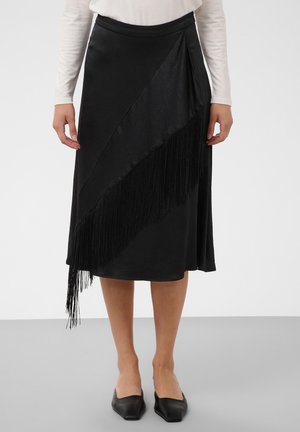 Femme portant une jupe midi noire avec un détail frangé diagonal sur le devant, associée à un haut blanc à manches longues et des ballerines noires pointues.
