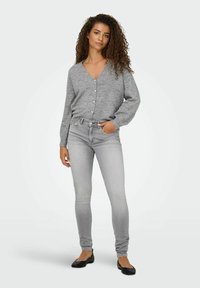 Cardigan en maille grise avec des boutons en perles, associé à un jean skinny gris clair et des ballerines noires. Cheveux bouclés portés lâchés.