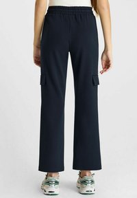 Vrouw in zwarte rechte cargo broek met elastische tailleband en wit-groene sneakers, gezien vanaf de achterkant op een effen achtergrond.