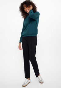 Teal sweatshirt met een ronde halslijn en ribgebreide manchetten, gecombineerd met zwarte broek. Model draagt witte en grijze sneakers en staat met een hand in haar haar.