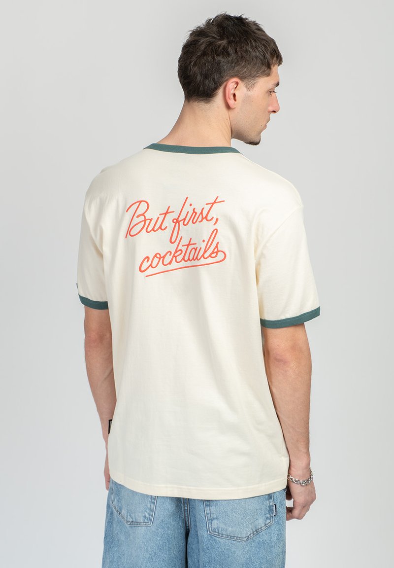 Άνδρας που φοράει κρεμ μπλουζάκι με πράσινο περίγραμμα και κόκκινο κείμενο «Αλλά πρώτα, κοκτέιλ» στην πλάτη, συνδυασμένο με ανοιχτόχρωμα τζιν σορτς.