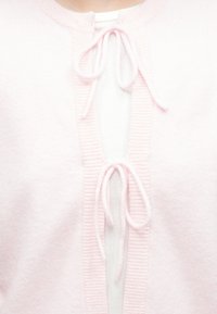 Zachte roze cardigan met ribgebreide randen en een striksluiting bij de hals, gelaagd over een witte textuurtop.