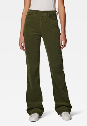 Osoba nosząca szerokie spodnie sztruksowe w oliwkowej zieleni z zapięciem na guziki i zamek błyskawiczny, w połączeniu z białymi niskimi trampkami.