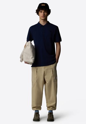 Young man wearing navy polo, beige cargo pants, green sneakers, black cap, holding a white tote bag, standing against a plain background.