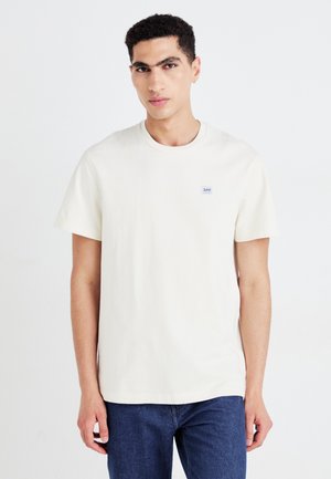 Kortermet kremfarget bomull T-skjorte med rund hals og lite blått logo-lapp på brystet. Kombinert med mørk denim jeans.