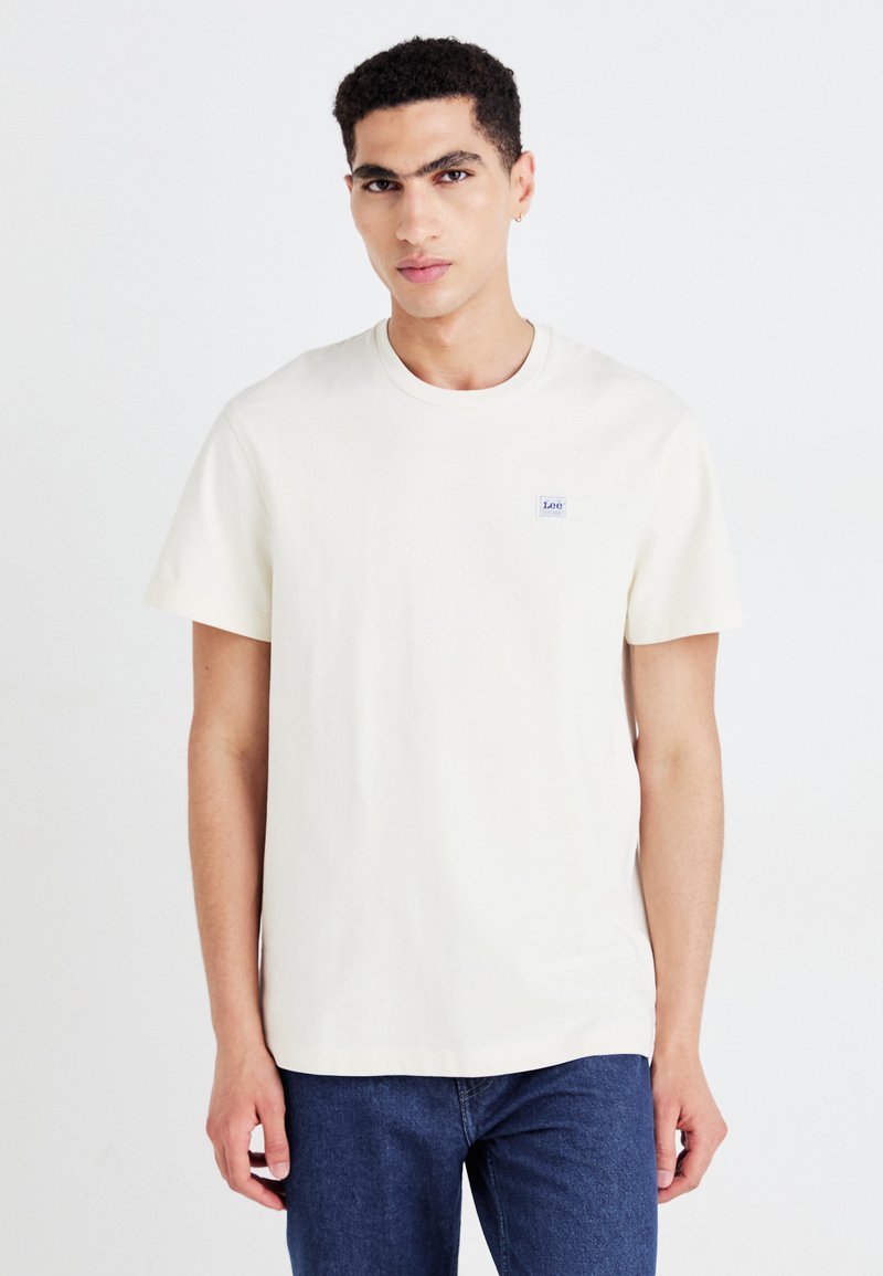 Korte mouwen crème katoenen T-shirt met een ronde halslijn en een klein blauw logo-opdruk op de borst. Gepaard met donkerdenim jeans.