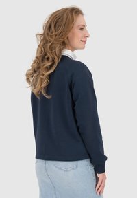 Felpa navy con colletto a girocollo, orlo a coste e maniche lunghe. Presenta una texture liscia. Abbinata a jeans azzurri chiari.