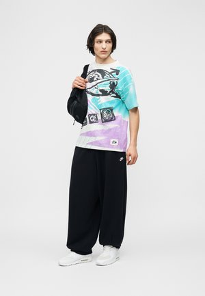 Νέος άνδρας που φοράει ένα μπλουζάκι σε πρασινομπλε και μωβ χρώμα με σχέδια, μαύρο παντελόνι φαρδύ, λευκά αθλητικά παπούτσια και κρατάει μια μαύρη τσάντα ώμου, στέκεται και κοιτάζει προς τα αριστερά.