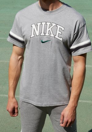 Person steht im Freien auf einem grünen Sportplatz und trägt ein graues Nike-T-Shirt sowie passende graue Shorts.