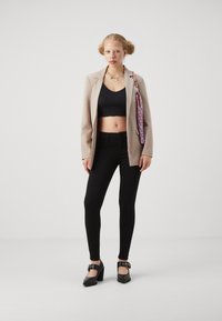 Beige blazer, black cropped top, black skinny pants, and black heeled shoes. Notable accessories include a colorful beaded necklace and handbag.