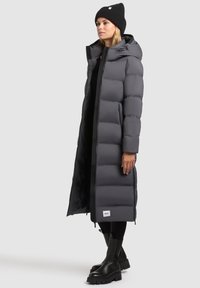 Lang grijs pufferjack met horizontale baffles, een capuchon en een rits aan de voorkant. Gedragen met een zwarte beanie en robuuste zwarte laarzen voor extra warmte.