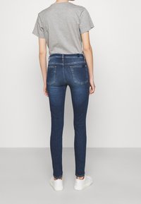 Jeans de denim azul ajustados con costuras en contraste, dos bolsillos traseros, combinados con una camiseta gris y zapatillas blancas.