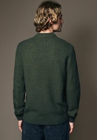 Dark green sweater with a textured grid pattern, ribbed neck and cuffs, and a relaxed fit. The back shows the design and stitching details.