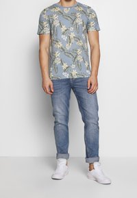 Krátký rukáv světle modrého trička s květinovým vzorem v žluté a zelené barvě, v kombinaci s vyhrnutými světle modrými džíny a bílými teniskami.