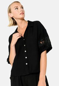 Camicia nera con maniche corte, tessuto con ricami floreali trasparenti sulle maniche, chiusura frontale con bottoni e un design classico del colletto.