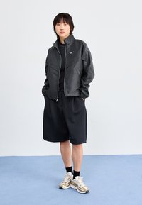 Casaco leve preto com gola alta, fecho zip, e punhos elásticos; combinado com calções pretos e sapatos de desporto com detalhes dourados.
