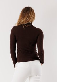 Maglietta marrone a maniche lunghe con design aderente e texture rigata, abbinata a pantaloni bianchi aderenti. La vista posteriore mette in risalto il colletto e la silhouette senza cuciture.