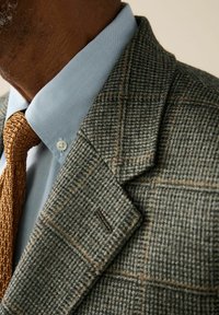 Man wearing a grey checkered blazer, light blue dress shirt, and textured brown knit tie, showing collar and upper chest area.