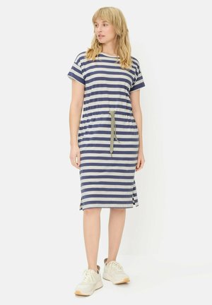 Kobieta stojąca w granatowo-białej sukience w paski do kolan, z krótkimi rękawami, wiązaną z przodu w talii, w białych trampkach.