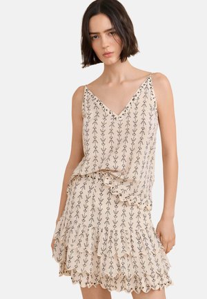 Vrouw met kort donker haar die een mouwloze beige jurk draagt met een zwart bladerenpatroon en een gelaagde geschulpte zoom tegen een witte achtergrond.