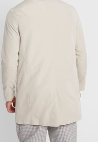 Kehopäiväinen, vaalea beige takki suoralla leikkuulla, jossa on takasauma ja sileä pinta, yhdistettynä ruudullisiin housuihin.