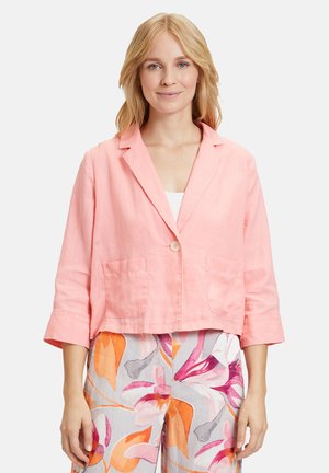Blazer cropped in lino rosa con due tasche frontali e chiusura con un bottone, indossato sopra pantaloni con motivo floreale.