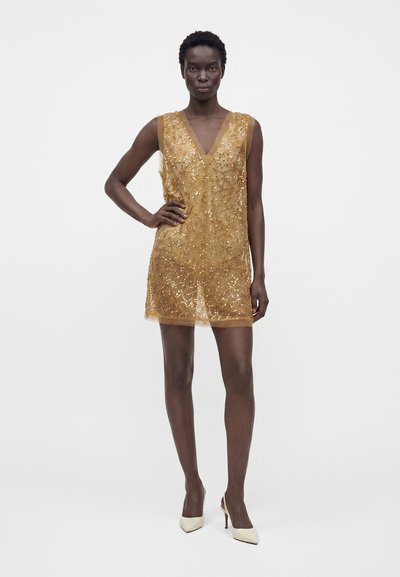 Femme à la peau foncée portant une mini-robe sans manches en sequins dorés et des talons hauts blancs, debout avec une main sur la hanche sur un fond uni.